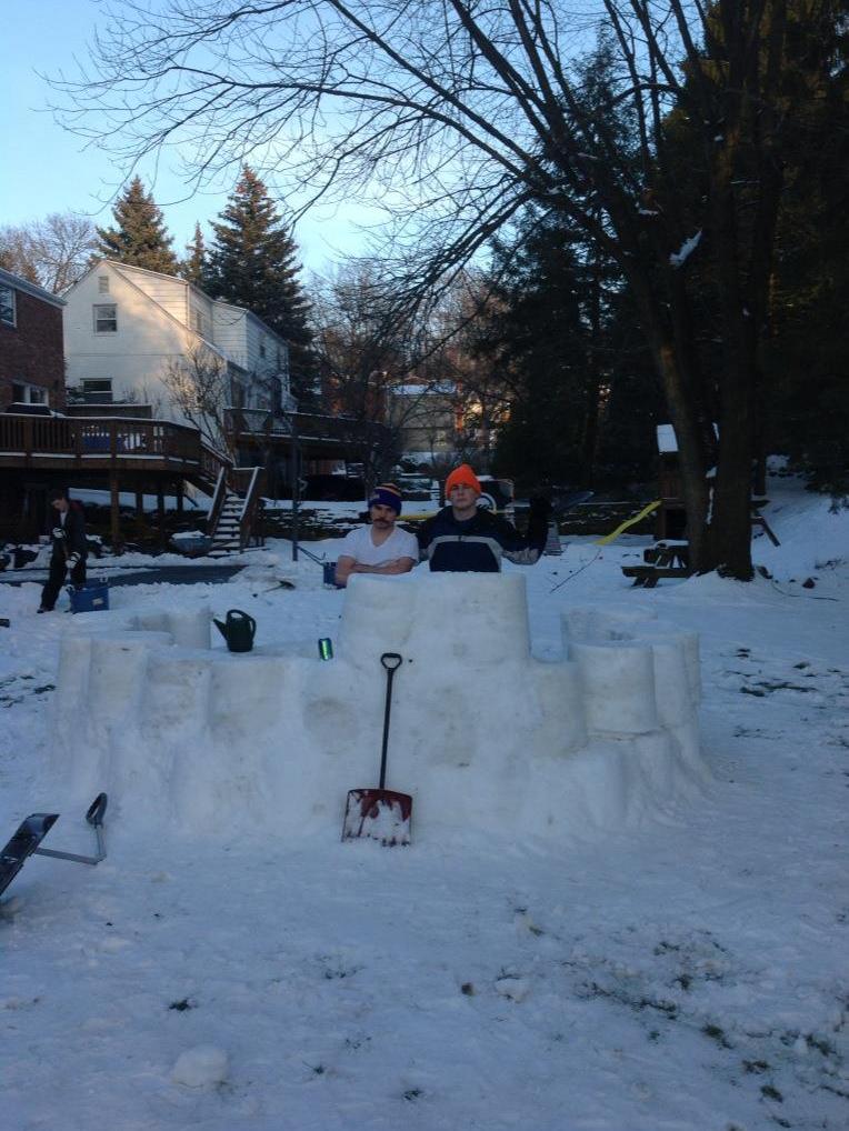 Two males standing behind a snow fort.