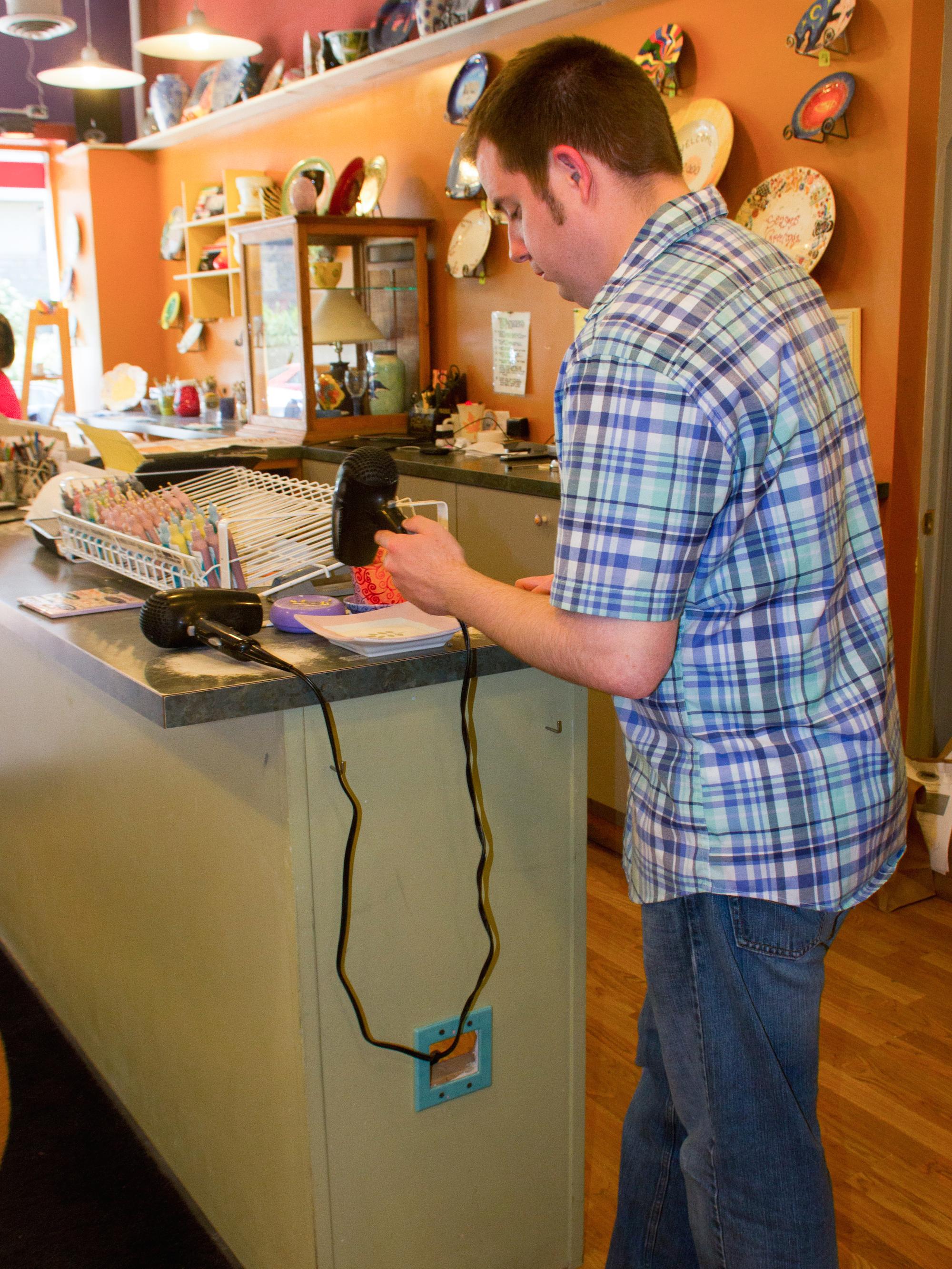 Jeremy Warner uses a hair dryer to dry his pottery at "Decorate your own bowl, mug, or plate at 2000˚ Degrees". 