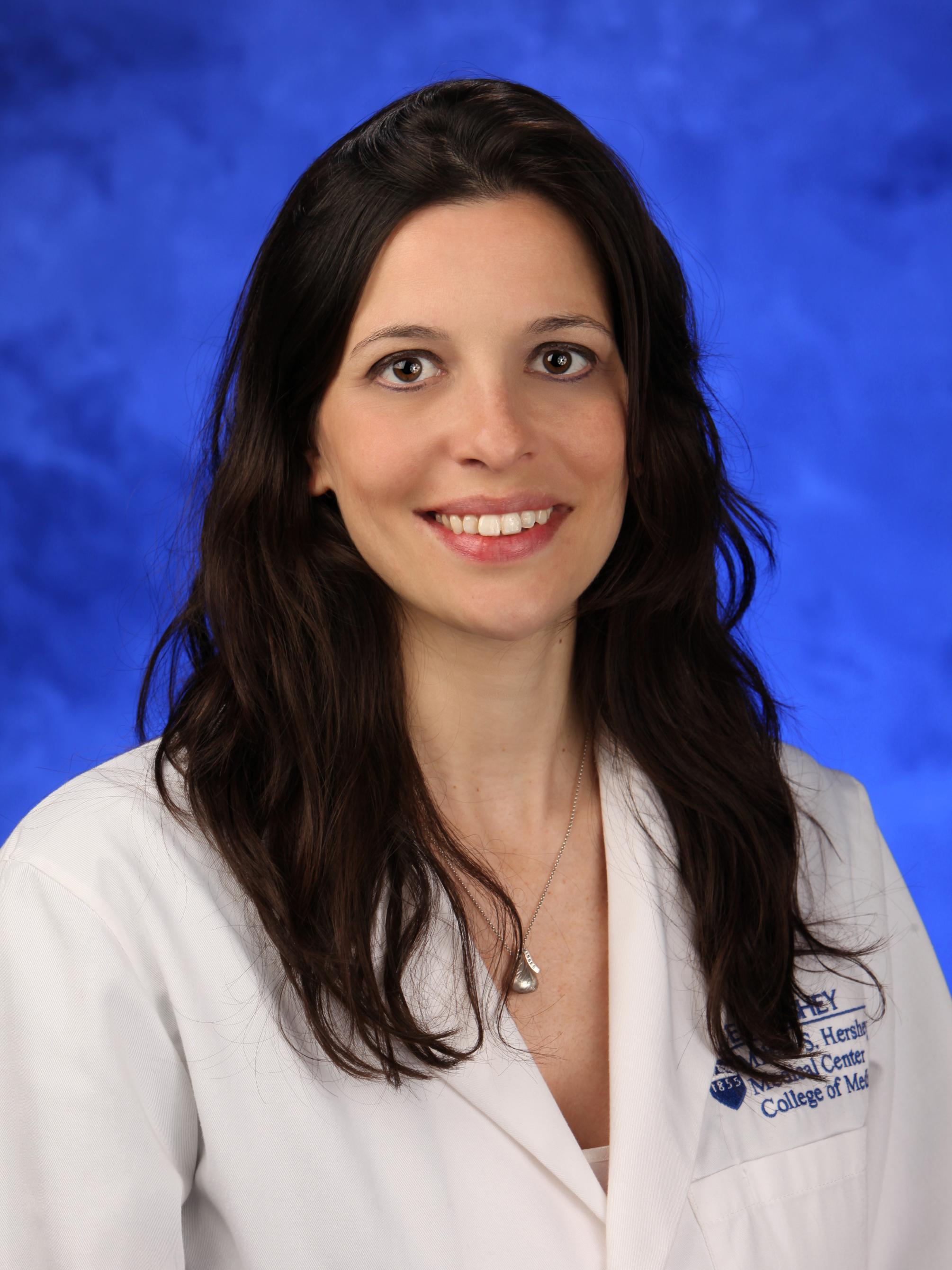 Dr. Bettina Aprile smiles into the camera, wearing a lab coat and collared shirt.