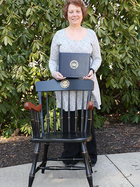 Woman standing behind chair and posing with certificate in front of green trees.
