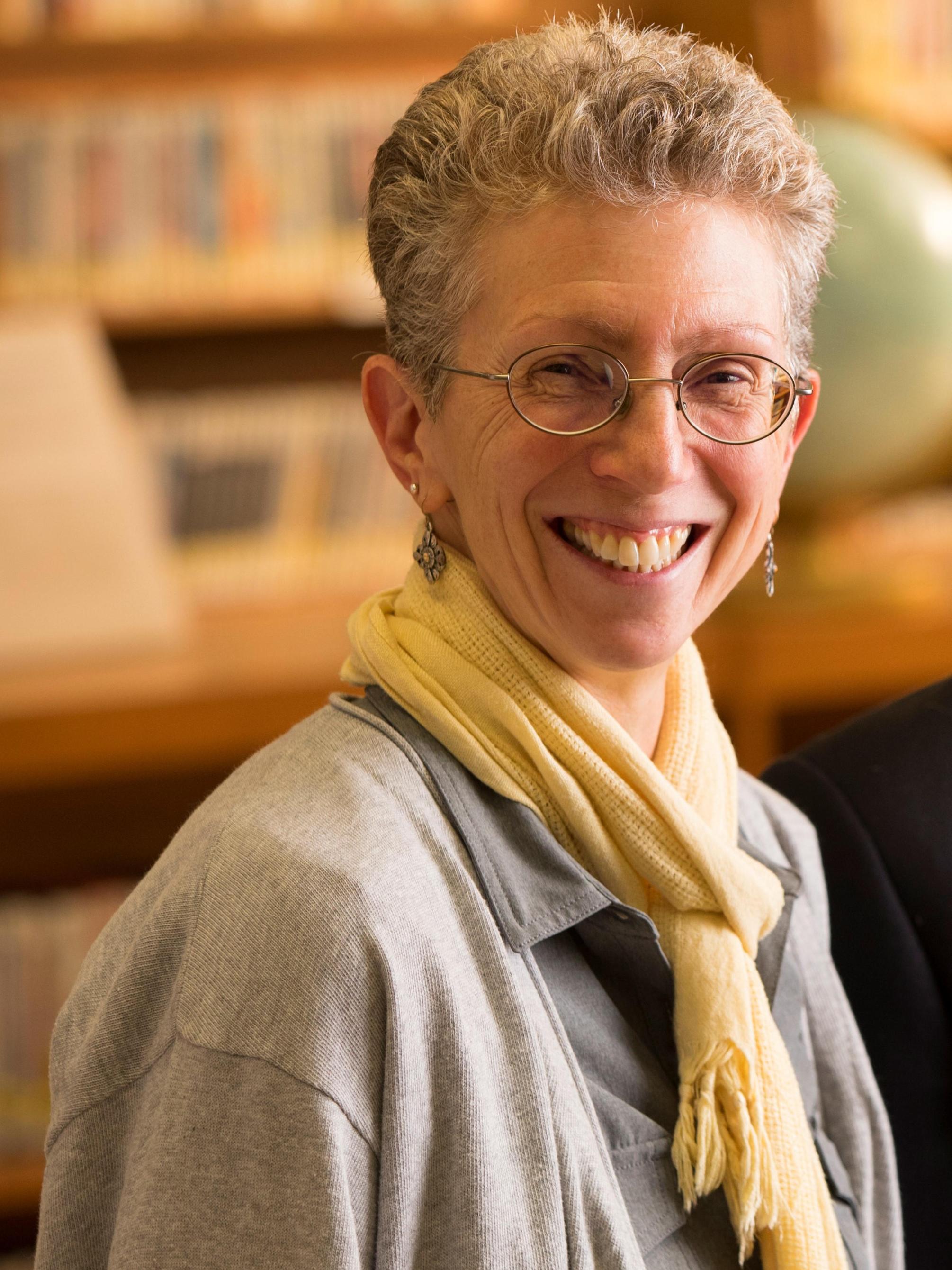 A woman with short hair and classes smiles for a photo in front of a bookshelf.