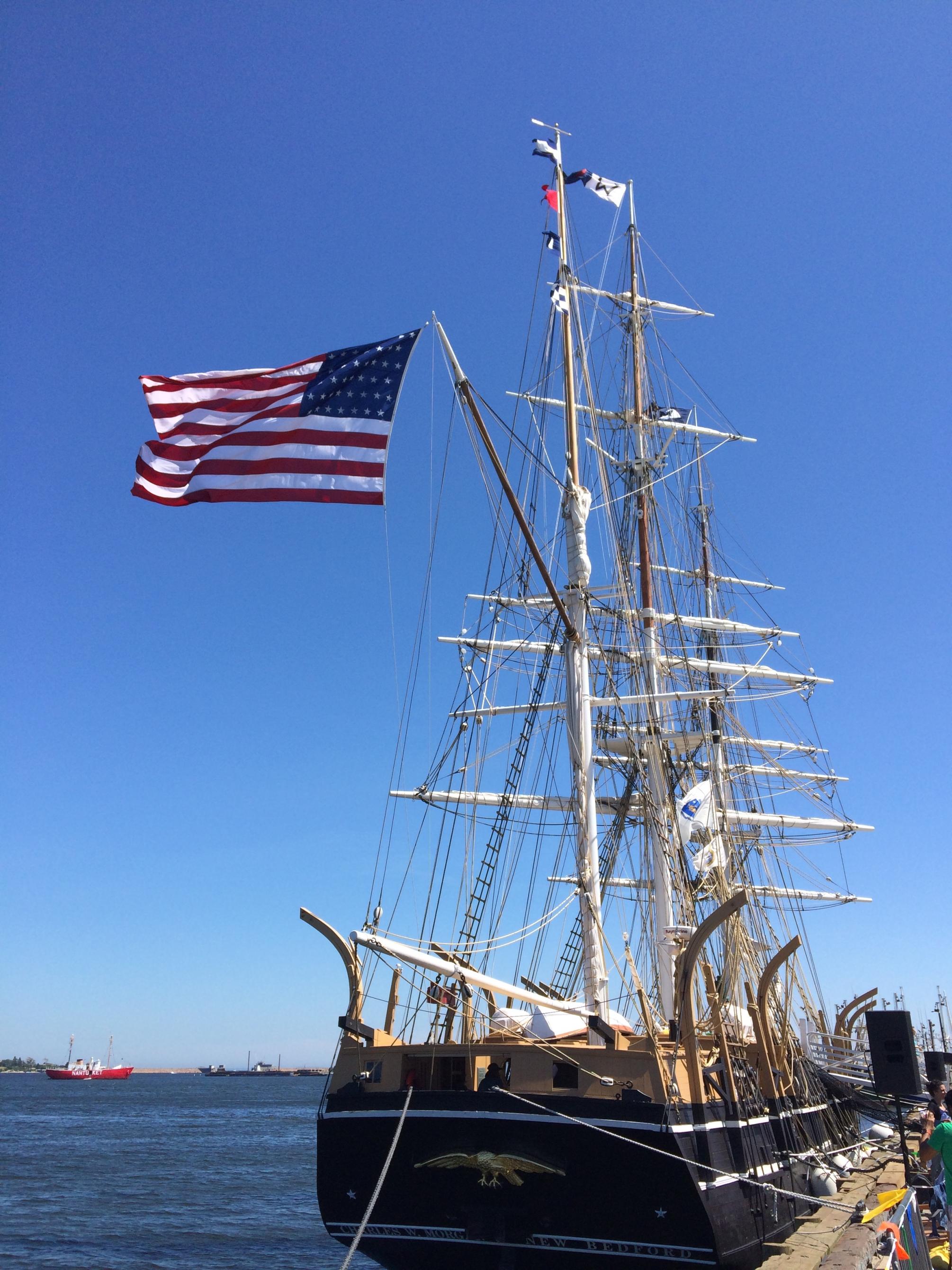A large wooden whaleship stands docked with the American flag flying in the wind. 
