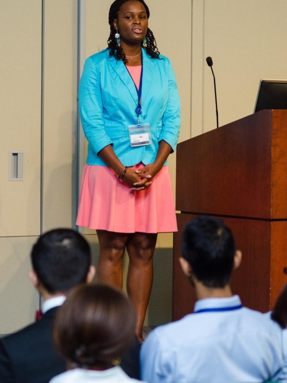 A woman stands in front of a small group speaking. To her left is a lectern with a microphone.
