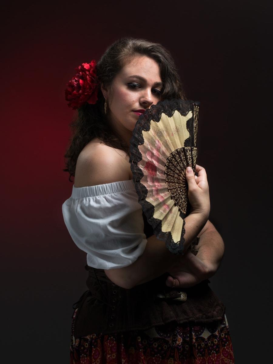 A woman looks at the camera as she holds a folding fan in front of her face.