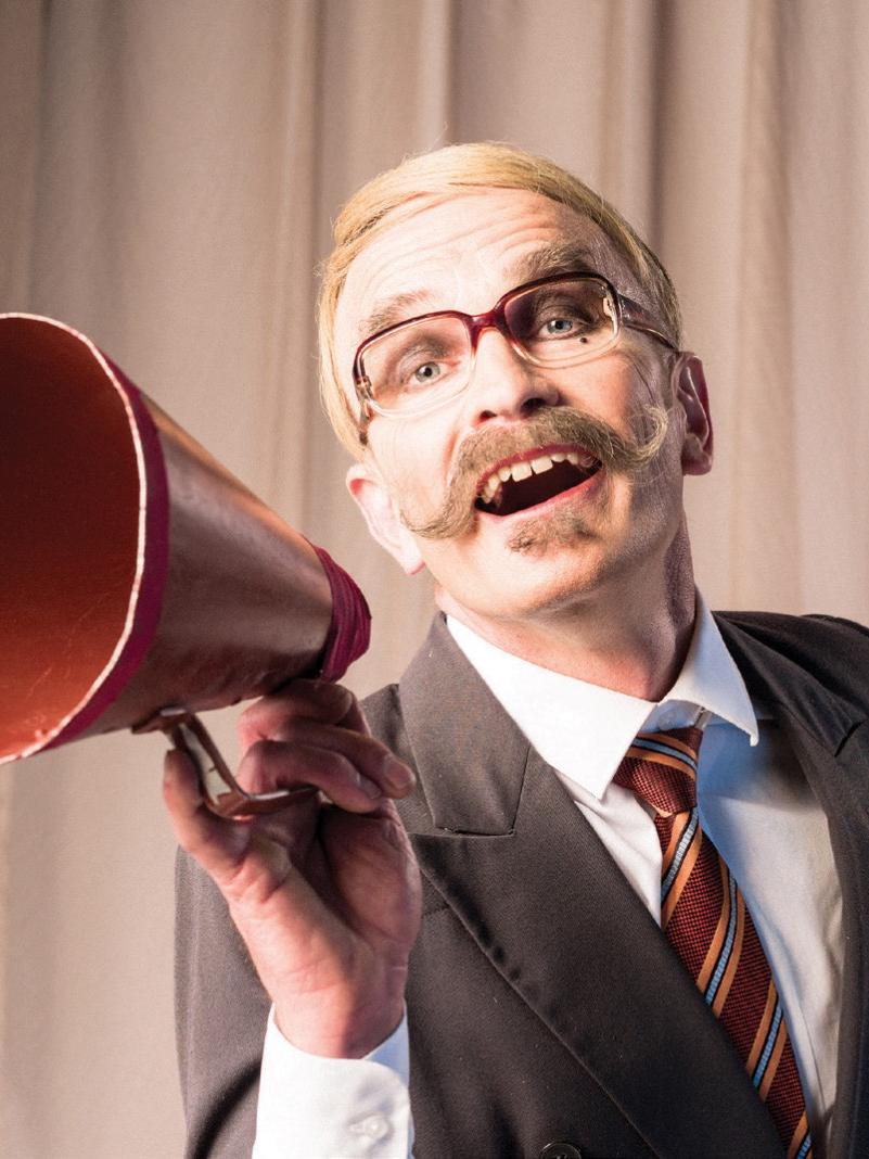 Circus Oz performer Matt Wilson, wearing a blonde wig, glasses and a suit, appears to speak into a megaphone.