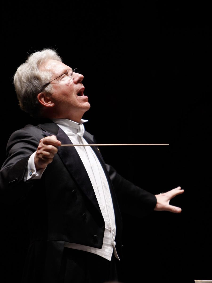 A man wearing a formal tuxdeo holds a conductor’s baton in his right hand while he looks upward.