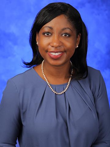 A woman with a blue blouse and a gold necklace poses and shoulder length black hair smiles in front of a blue background.
