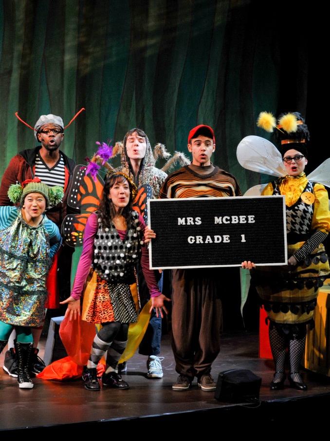 Actors dressed in street clothes to resemble insects stand in a huddle while a character dressed like a worm holds a sign.