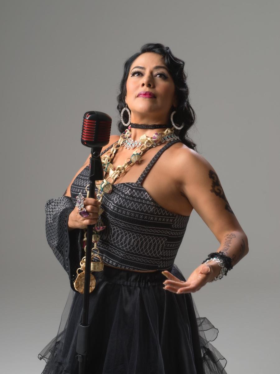 Lila Downs, wearing large baubles around her neck and on her fingers, holds a microphone in her right hand and looks up to the left.