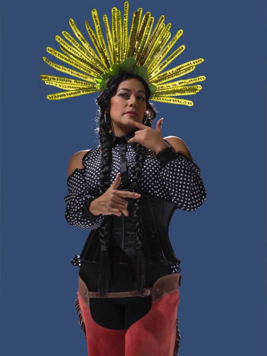 A woman wearing a feathered headdress and Western-style chaps makes gestures with her hands.
