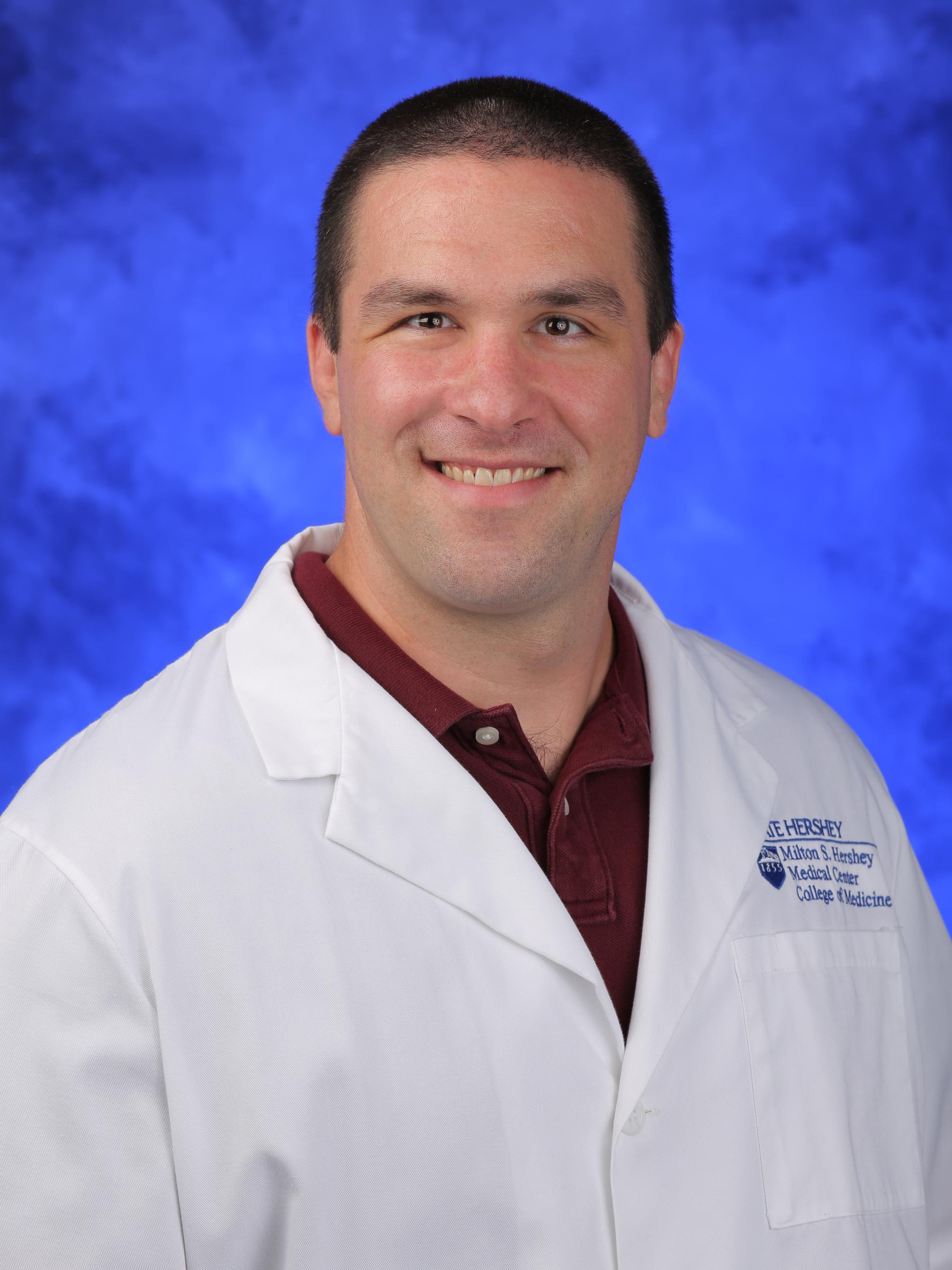 Anthony Dambro smiles into the camera, wearing a lab coat and collared shirt.