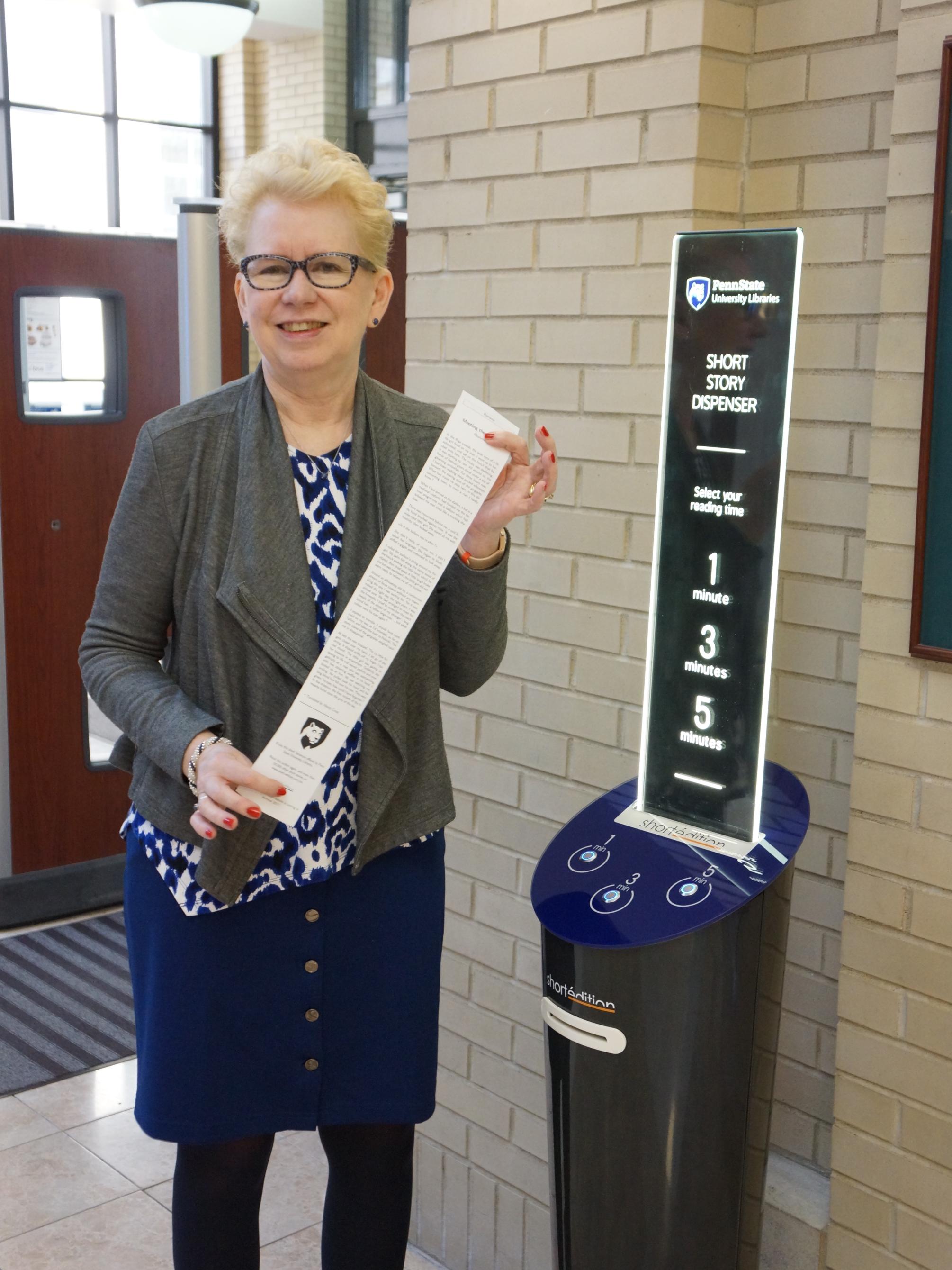 vertical image of smiling woman standing next to blue short story dispenser inside Penn State library entrance and holding long string of paper with short story printed on it from dispenser