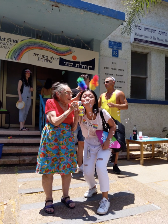 Young woman in a white t-shirt, white jeans and white sneakers blows bubbles with an older woman wearing a floral skirt and red t-shirt on the street.