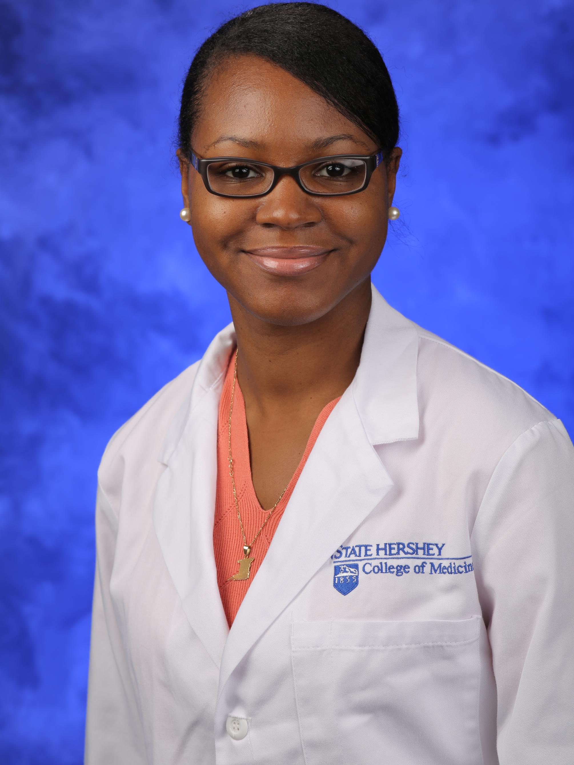 Annyella Douglas poses in front of a blue backdrop, wearing a peach-colored shirt with a white coat bearing the Penn State College of Medicine logo.