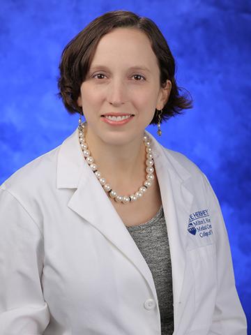 A woman with short brown hair wears a white doctors coat with a gray shirt underneath. A large pearl necklace and dangling pearl earrings are also being work. She is smiling in front of a blue background.
