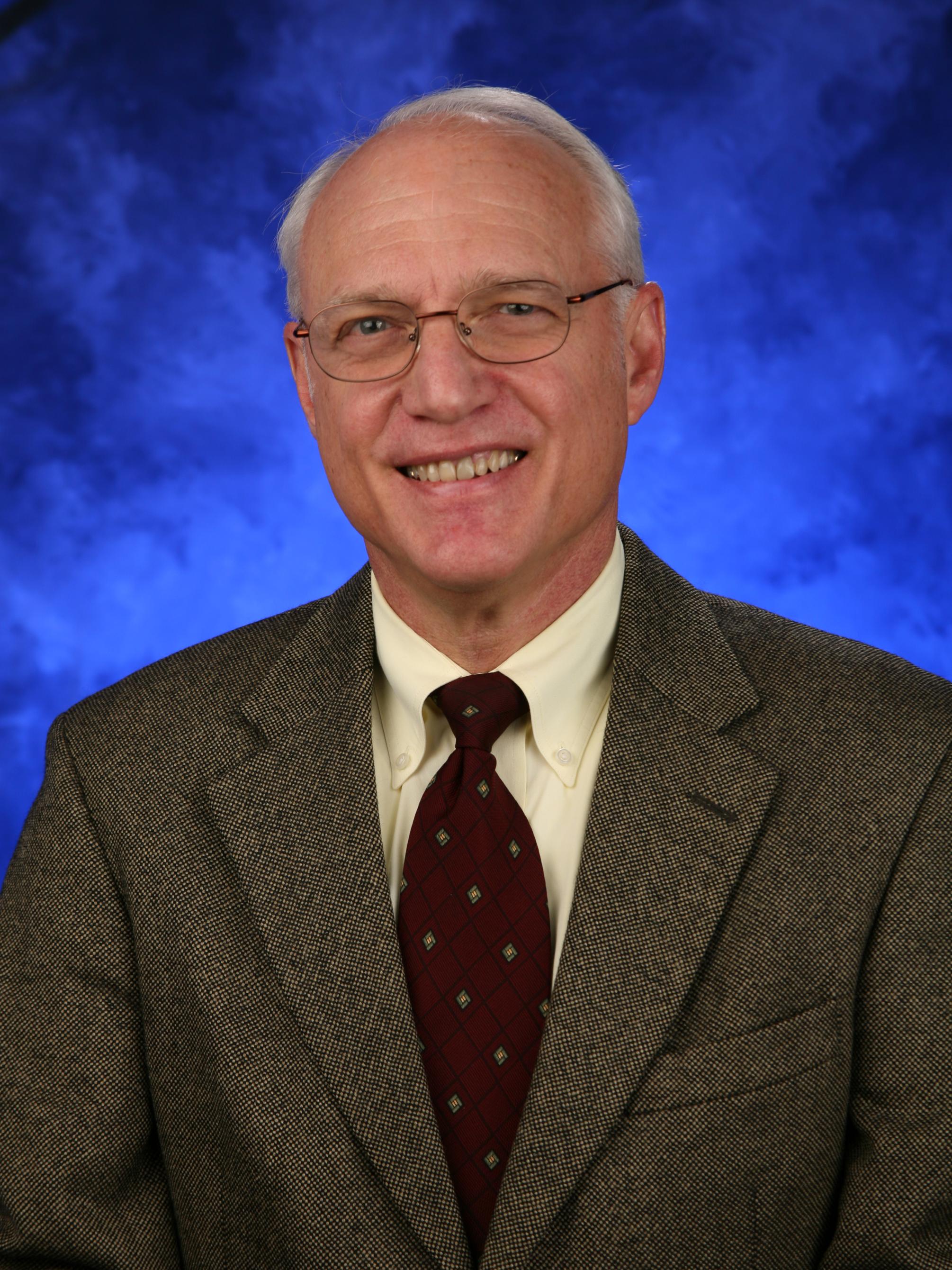 Mark Faulkner poses for a professional headshot, wearing a suit and tie.