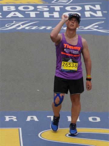 Tim Harner stands at the finish line of the Boston Marathon, which is marked on the street. He looks upward with his right hand raised to the sky.