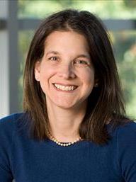 A woman with dark brown shoulder length hair wears a pearl necklace and blue shirt. She smiles and is front of a window with green shrubbery blurred.