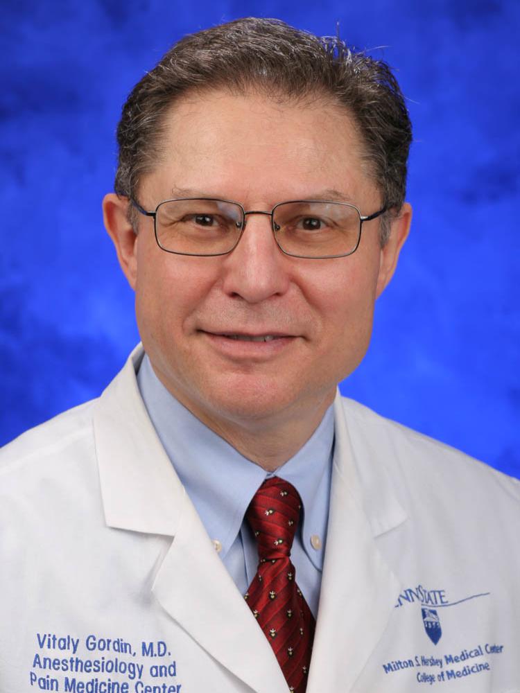 Dr. Vitaly Gordin smiles into the camera, wearing a lab coat, collared shirt and tie.