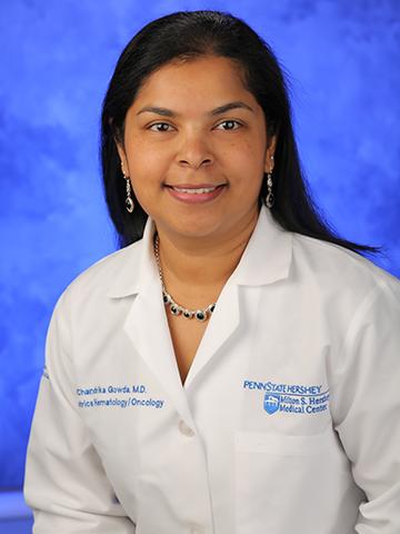 A woman with long black hair wears a white doctor coat with a silver and black necklace and earrings. She is smiling in front of a blue background.