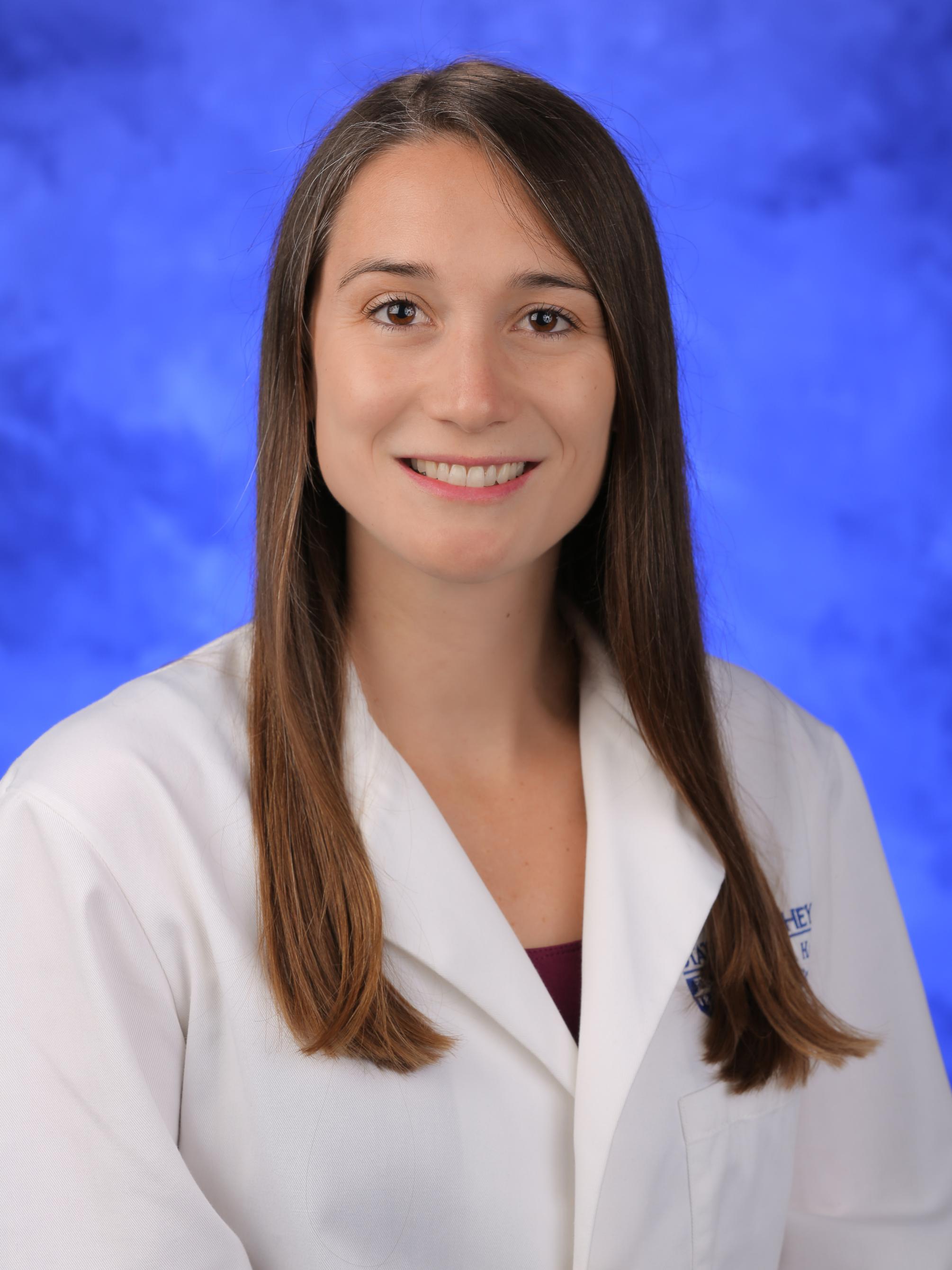 A woman with long brown hair smiles in front of a blue background. She is wearing a white doctor coat.