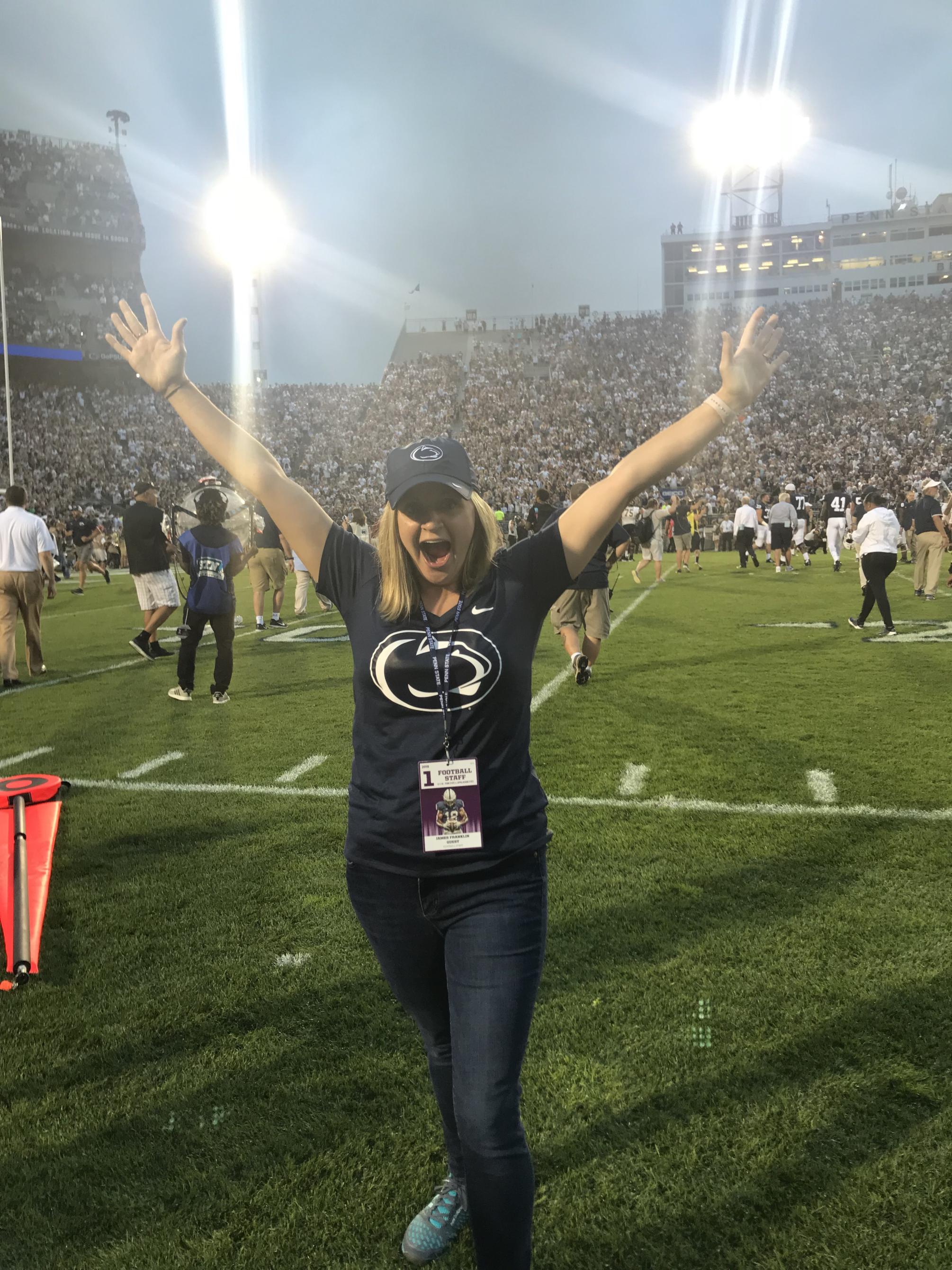 Woman stands on the sidelines of a football field with her hands in the air