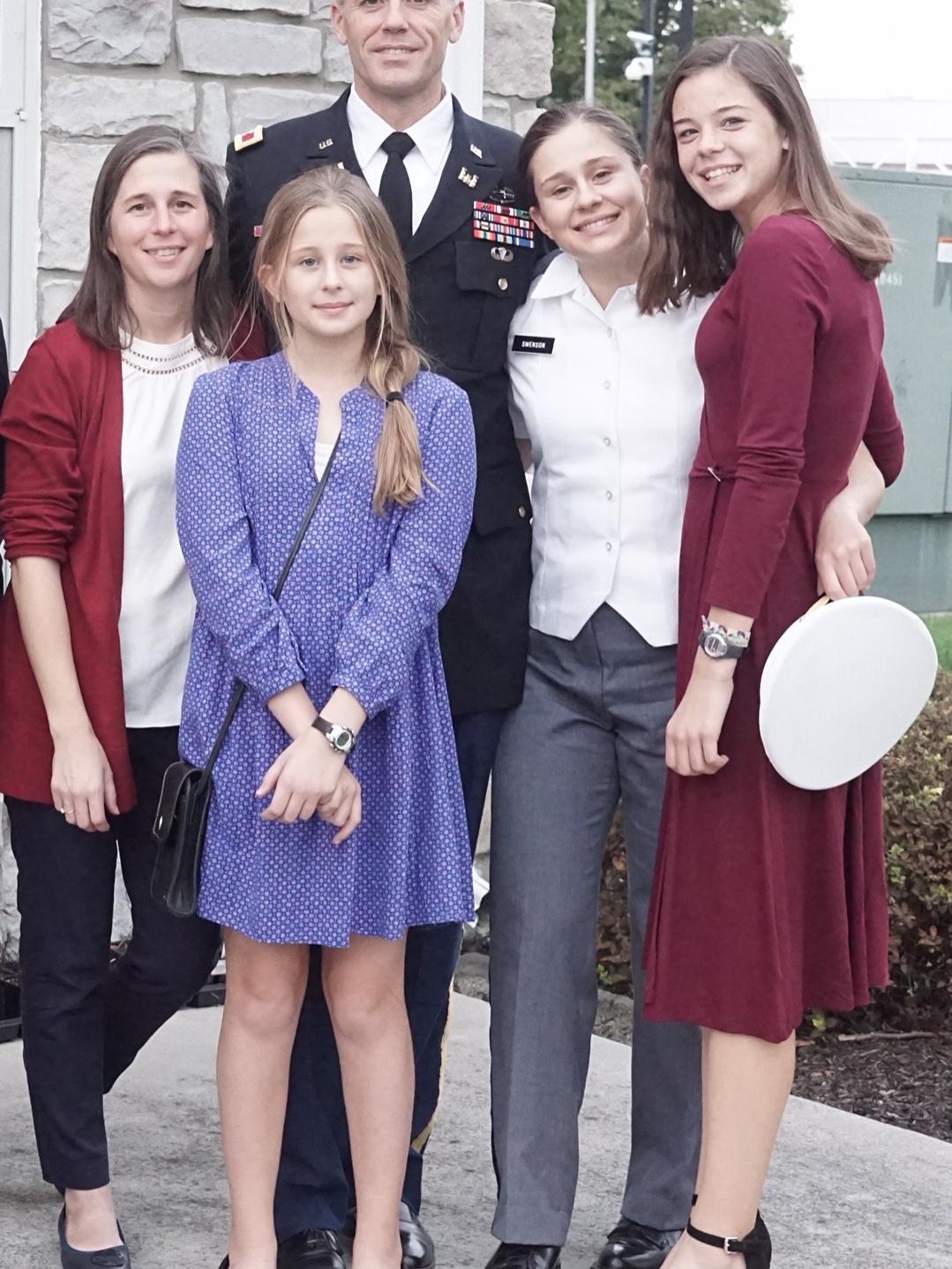 Man dressed in a military suit stands on a concrete porch with his wife and three young daughters.