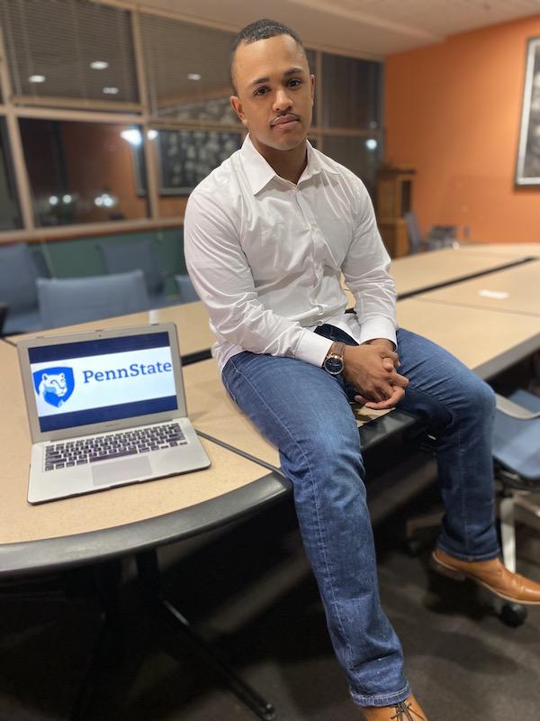 Man in a white long sleeve shirt and blue jeans sits on a table next to a computer with the Penn State logo on the screen.
