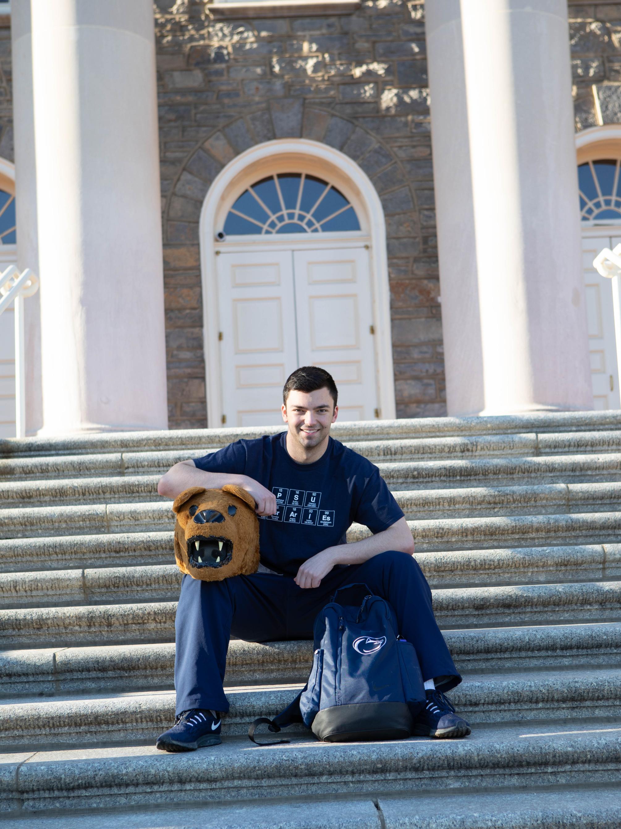 Zach Sowa sits on the steps of Old Main while holding the Nittany Lion mascot head.