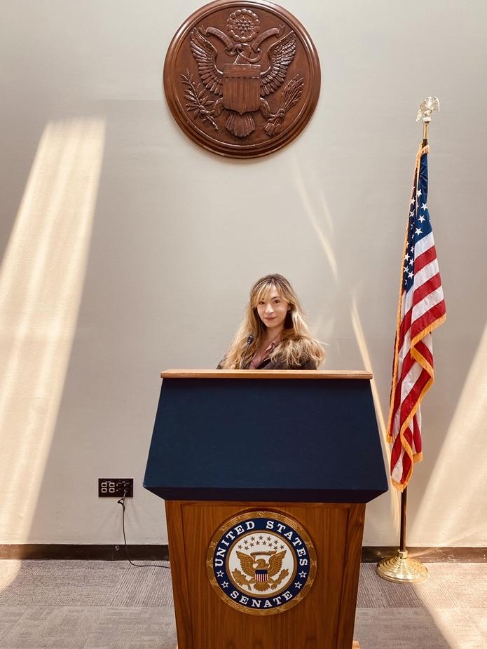 An image of student-intern Corrine Ellis standing at a podium with a U.S. Senate seal.