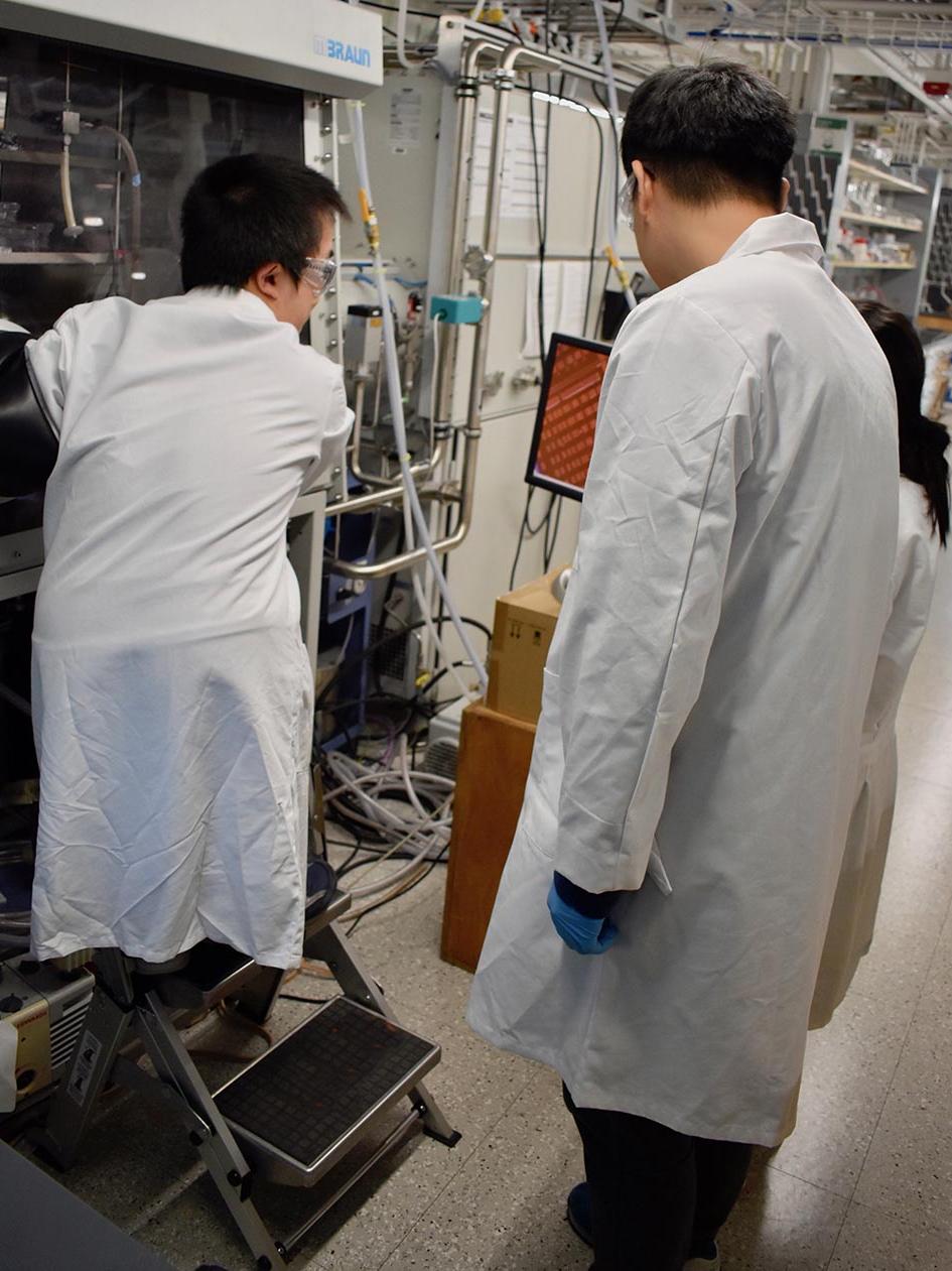 A man of short stature stands on a foot stool to work in a lab glove box while two others look on.