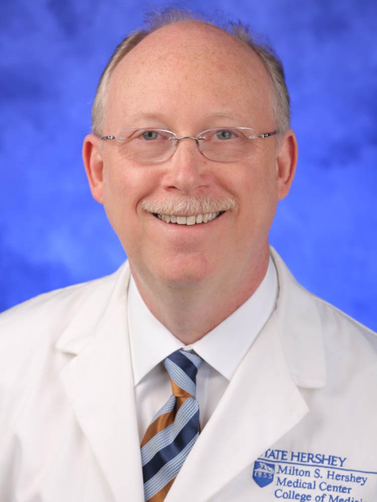 Dr. Jeffrey R. Kaiser wears a white coat and striped tie, posing in front of a blue, slightly textured backdrop.