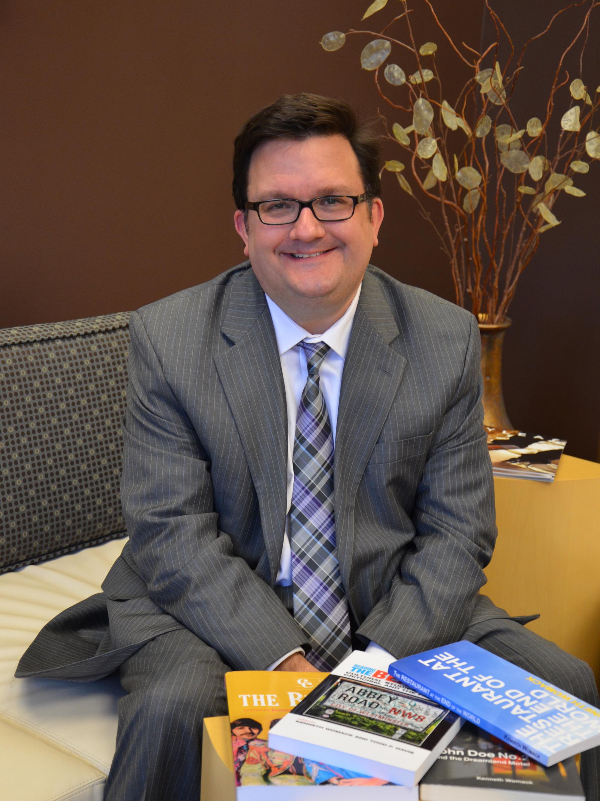 Kenneth Womack, man in suit seated on sofa in front of table of books he has authored