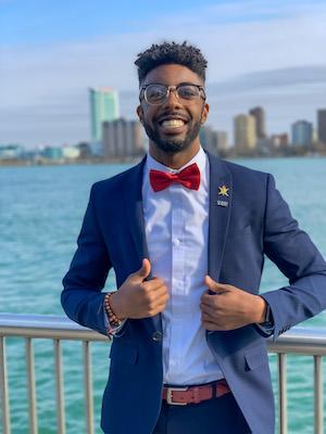 Kriston Ramdass in  suit and bowtie standing in front of body of water in a city, smiling and posing for photo