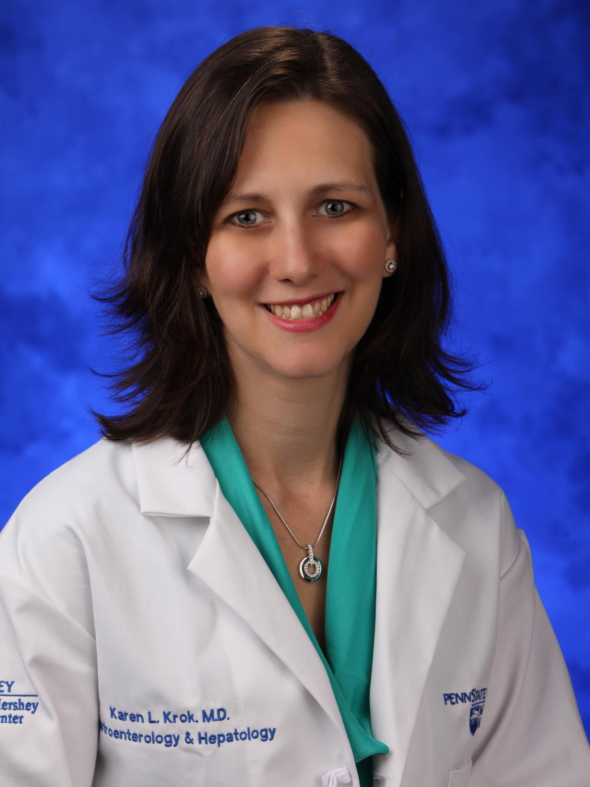 Dr. Karen Krok poses for a photo wearing a white physician's coat, in front of a blue lightly textured backdrop.