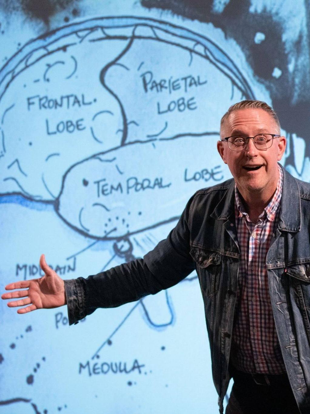 A man wearing a denim jacket stands in front of an illustration of a brain that identifies the various sections of the brain and their function