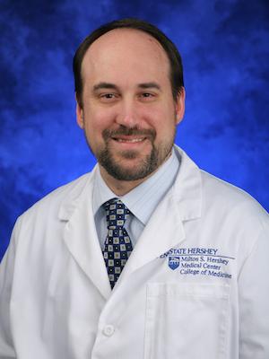 A portrait of a man in a tie and white coat against a blue background.