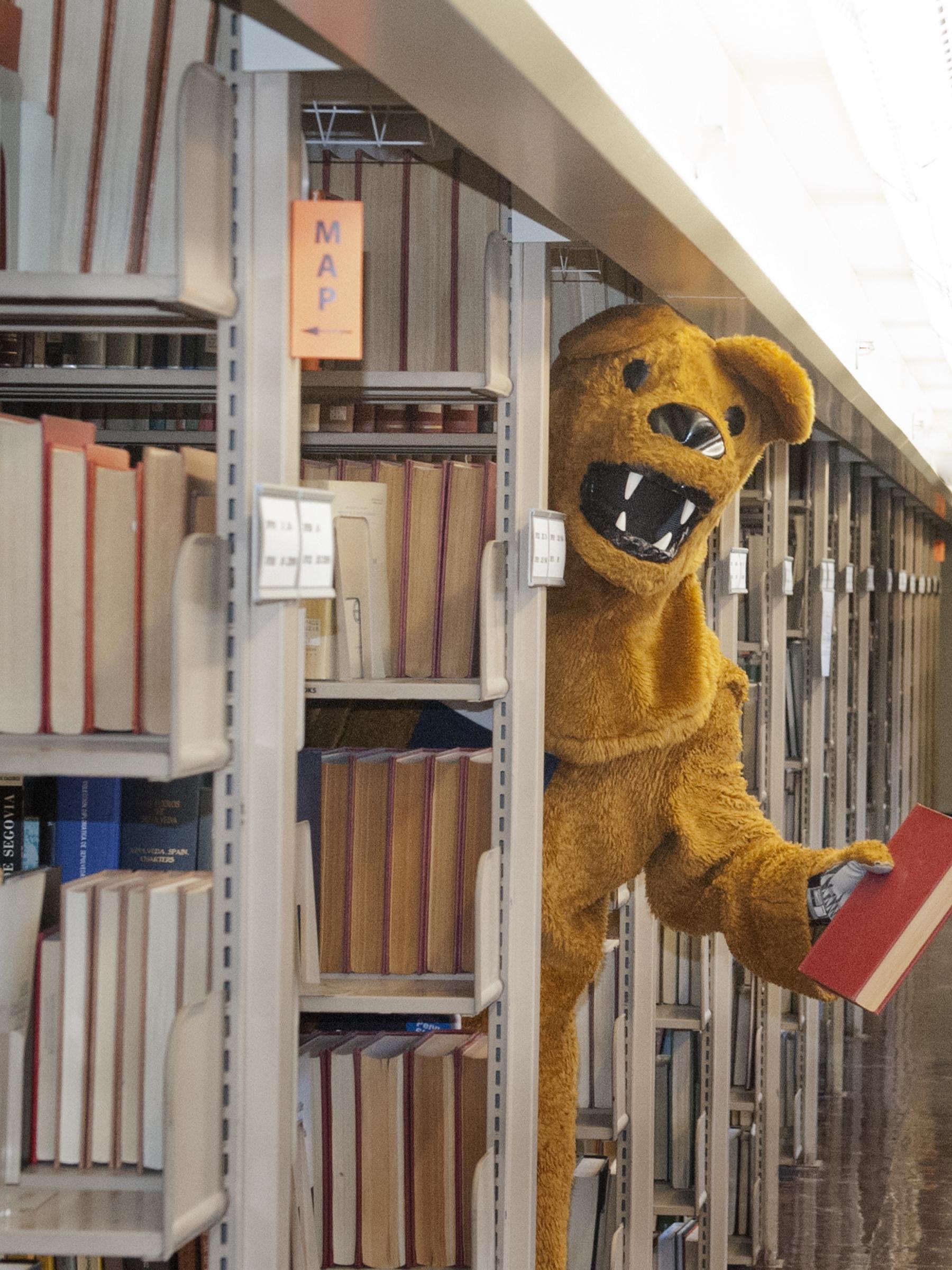 Nittany Lion mascot standing among library stacks bookshelves holding book