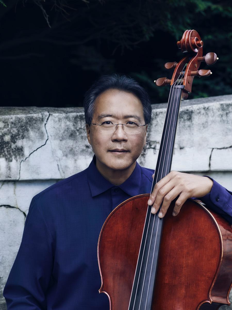 A man looks straight at the camera while he holds his cello upright and with his left hand on the neck.