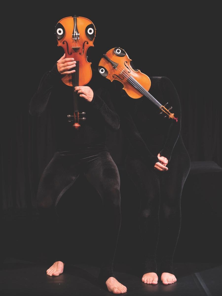 -- Two dancers hold violins decorated as faces in front of their heads.