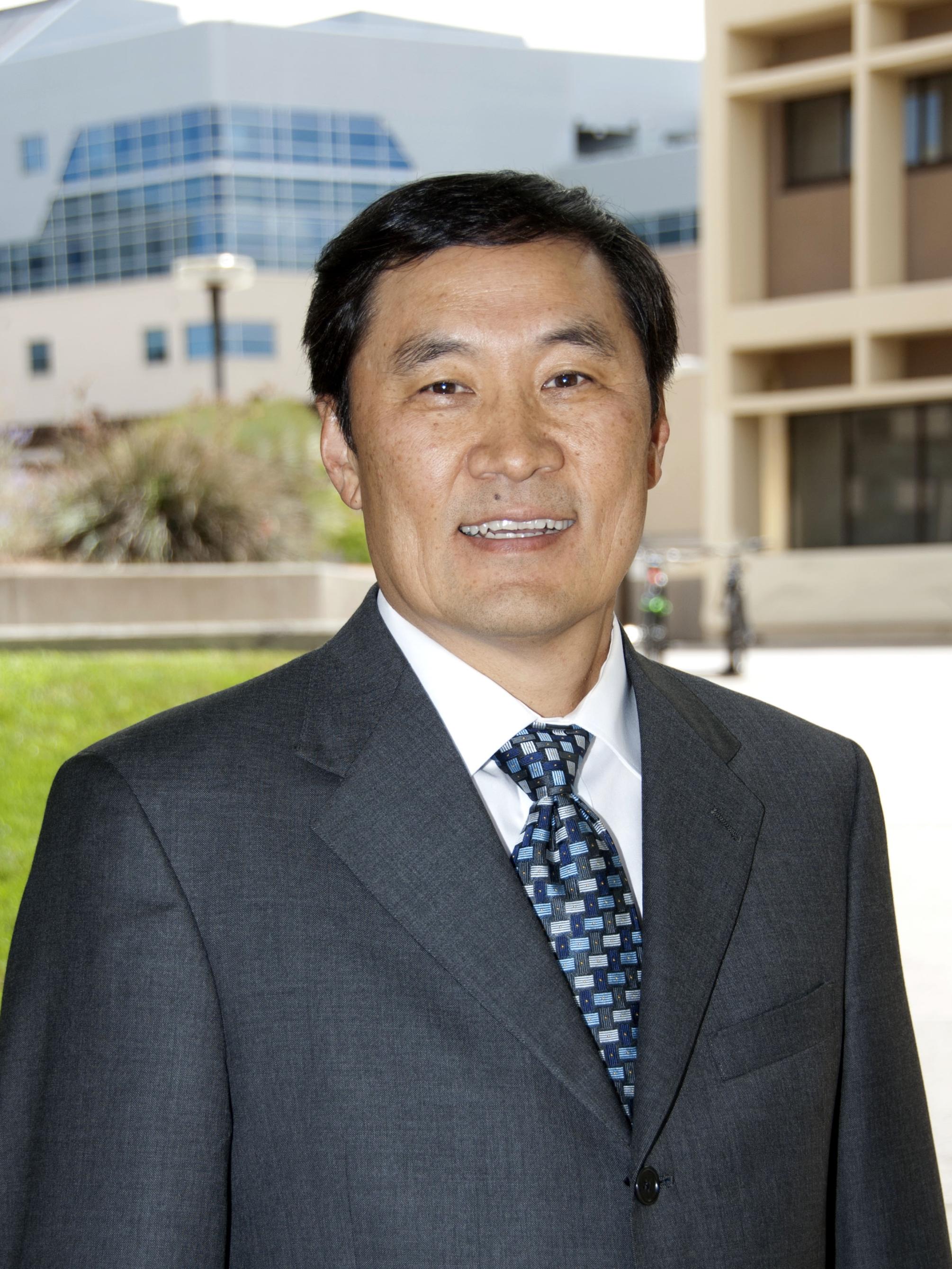 Dr. Thomas Y. Ma wears a suit, posing outdoors in front of a green lawn and two modern academic buildings.
