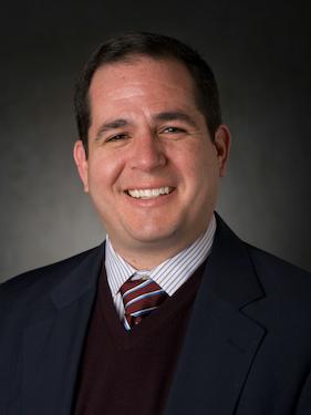 A portrait of a white man in a dark sweater and purple tie. 