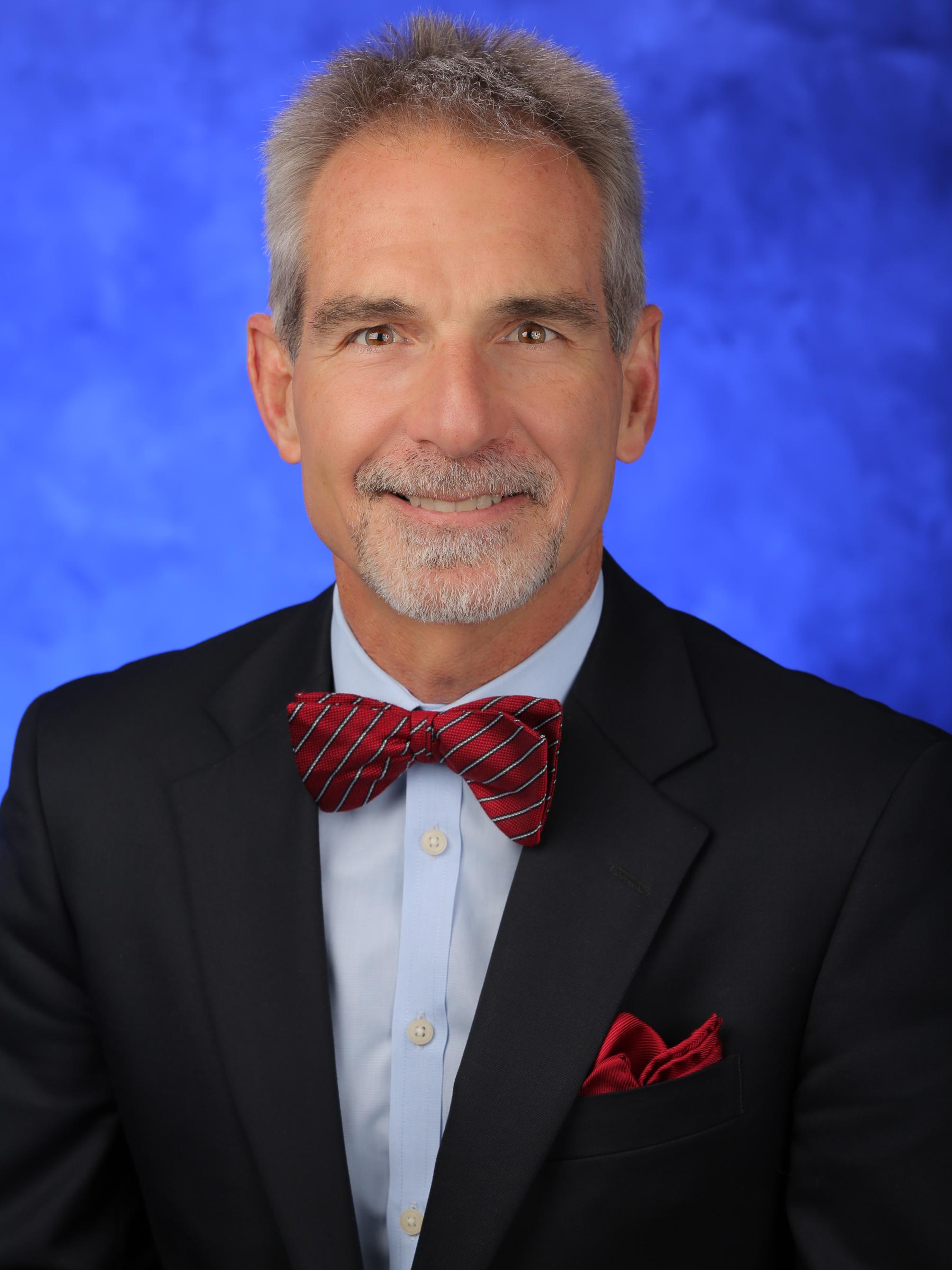 Dr. Berend Mets poses for a photo in a dark suit with a red bowtie, in front of a blue background.