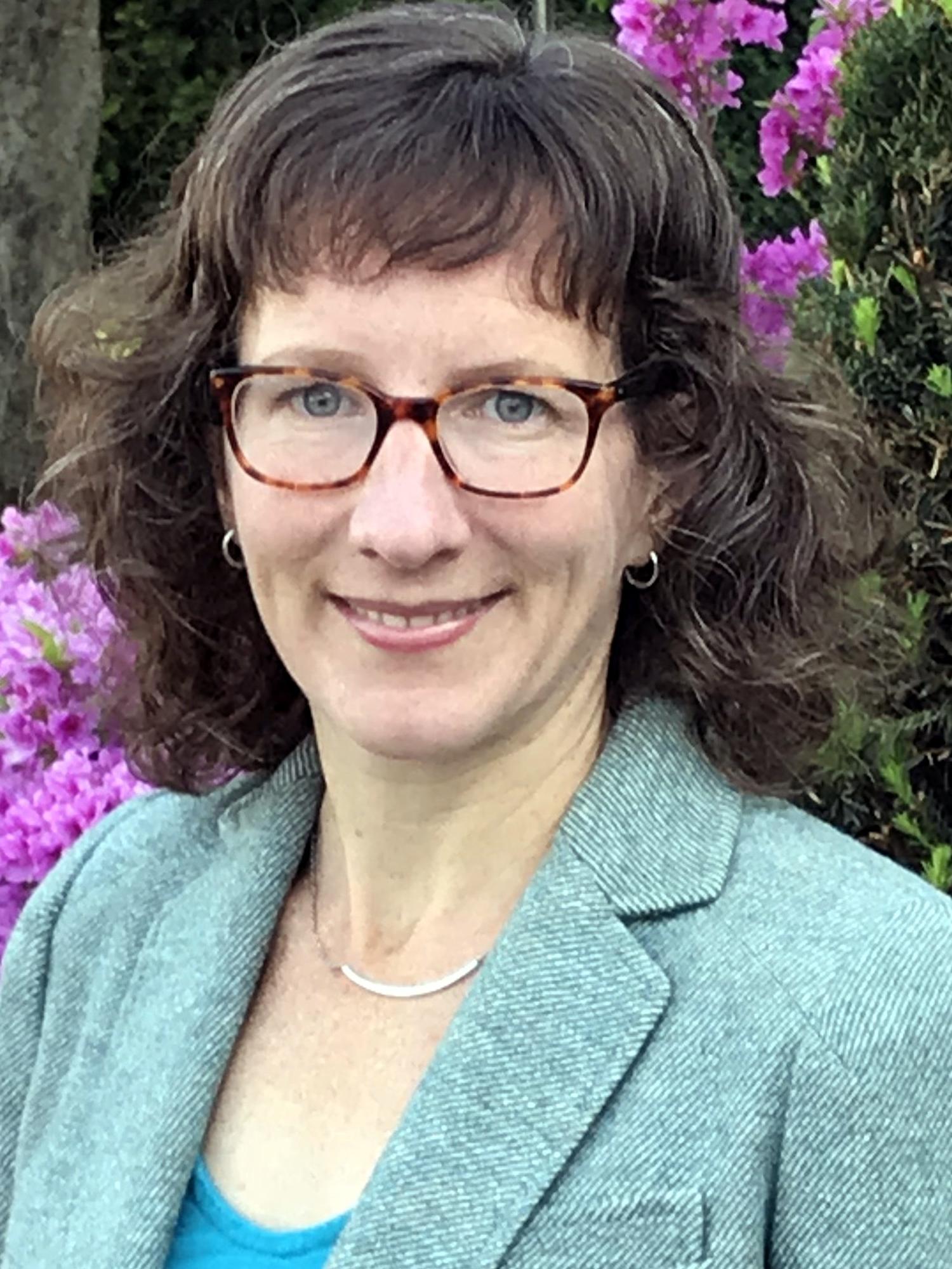 photo portrait of Tara Marray, woman wearing glasses and green blazer standing in front of purple flowers