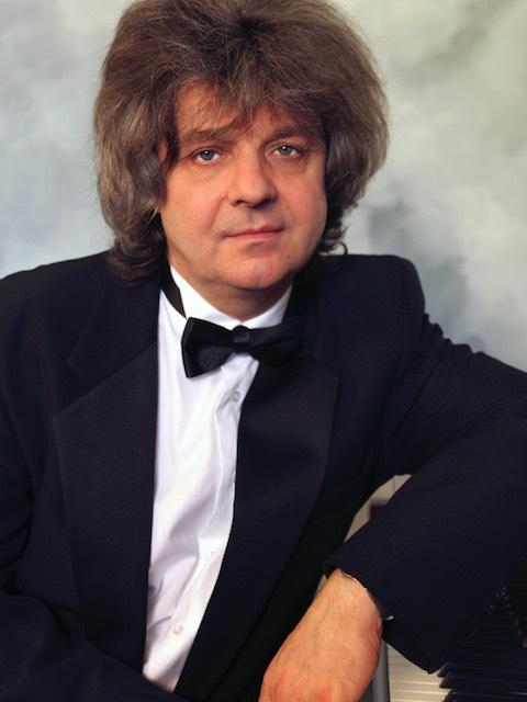 A man dressed in a tuxedo leans against a piano.