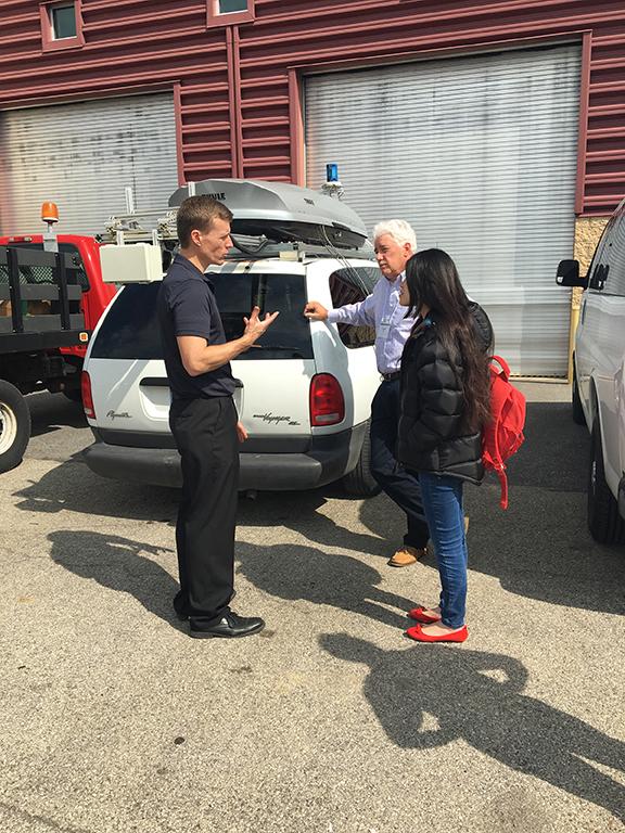 A man speaking to another man and woman while standing in front of a vehicle.