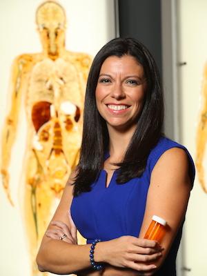 Woman in business professional attire holding a prescription bottle stands in front of an image of a human body and smiles for photo
