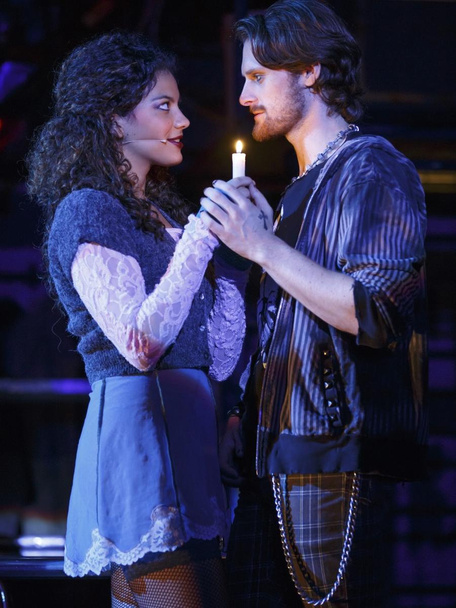 A young man and woman stand facing each other and jointly hold a candle in a scene from “Rent.” 