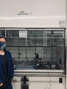student in lab attire poses next to a fume hood in an engineering lab. 