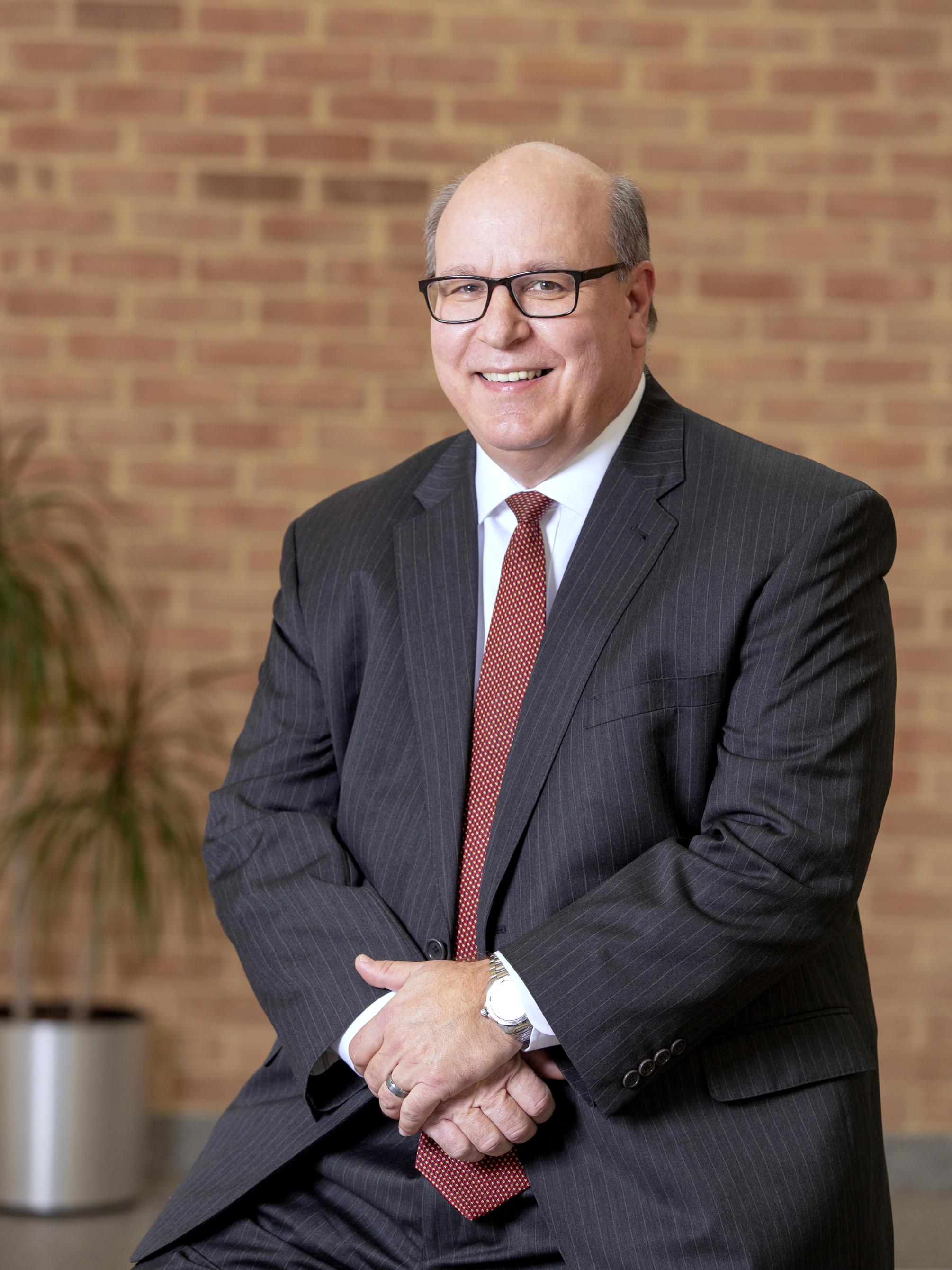 Steve Massini, wearing a suit, clasps his hands in front of a brick wall. He is flanked by a potted plant.